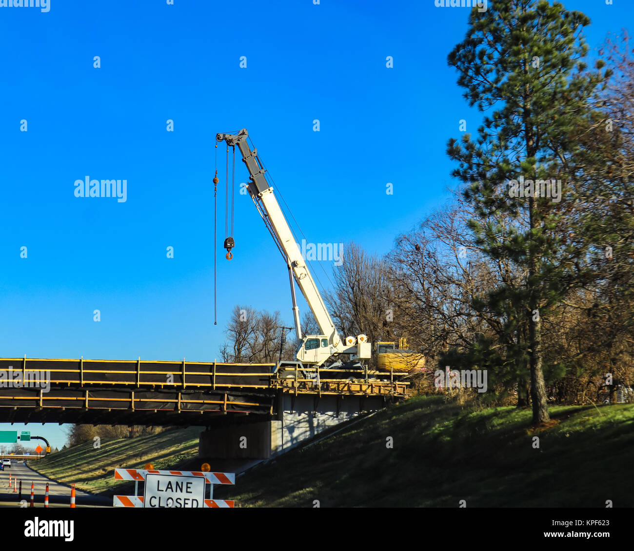 Grande grue mobile blanc travaillant sur un pont au-dessus de l'autoroute avec lane fermée par grand pin arbre et ciel très bleu Banque D'Images