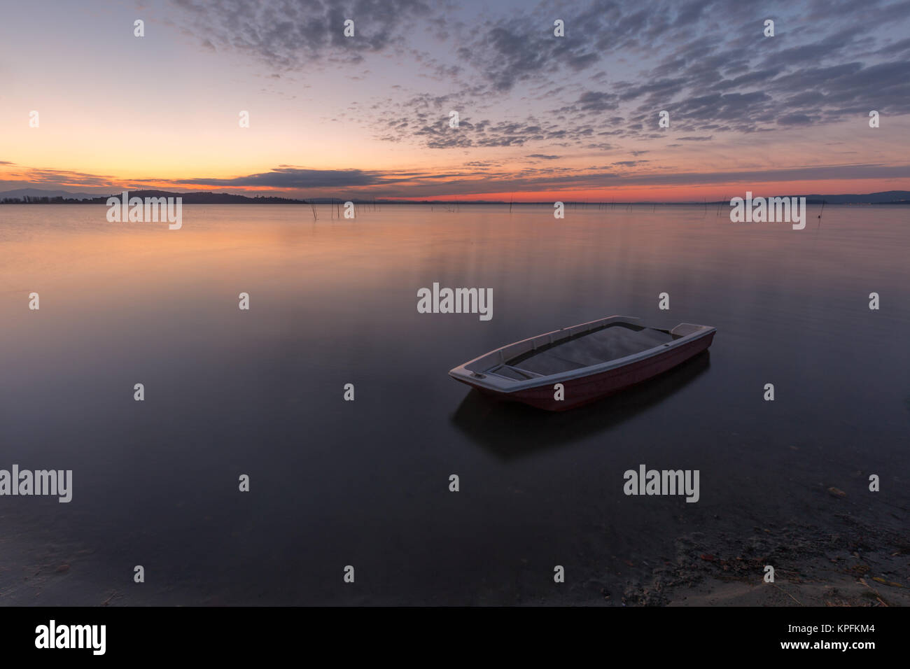 Un petit bateau de pêche dans le milieu de l'eau parfaitement immobile au coucher du soleil, avec le maquereau nuages ciel (lac Trasimène, Ombrie, Italie) Banque D'Images