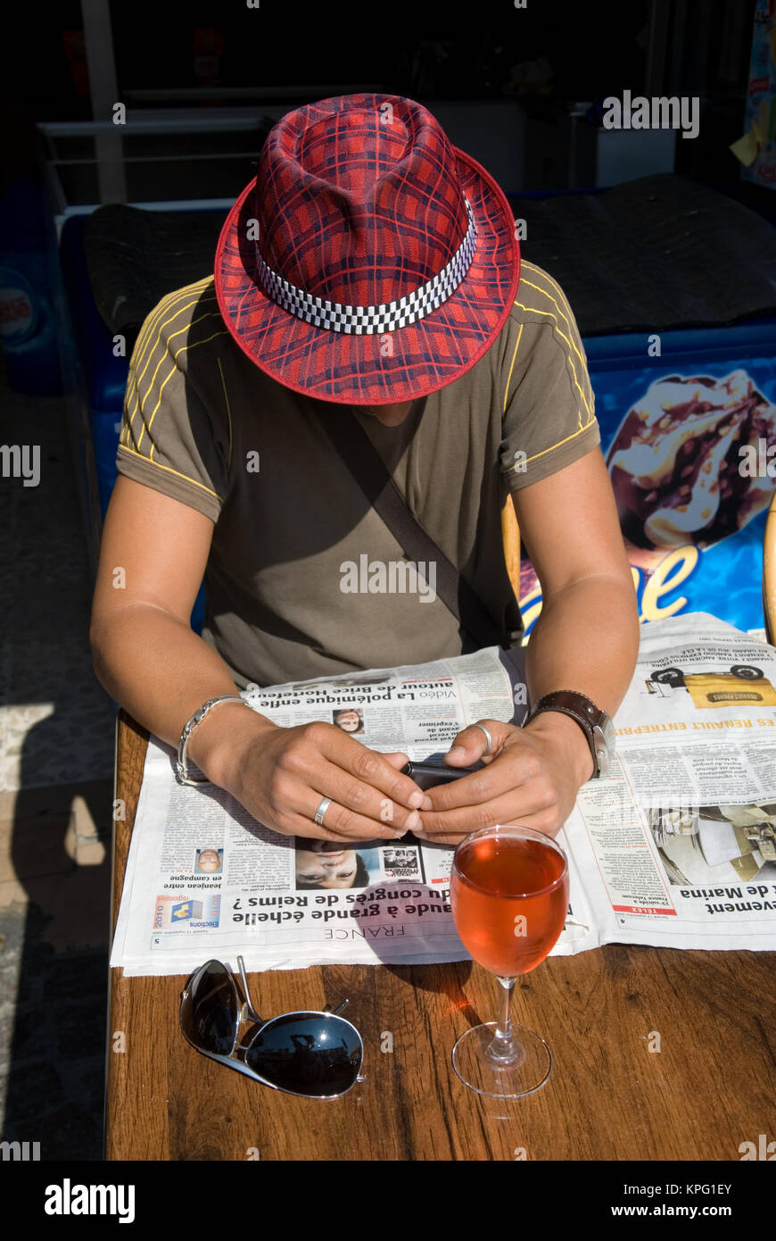 Jeune homme portant un chapeau lit un journal français à une table sur une  terrasse de café en France. Un verre de rosé frais et lunettes de s'asseoir  en premier plan Photo