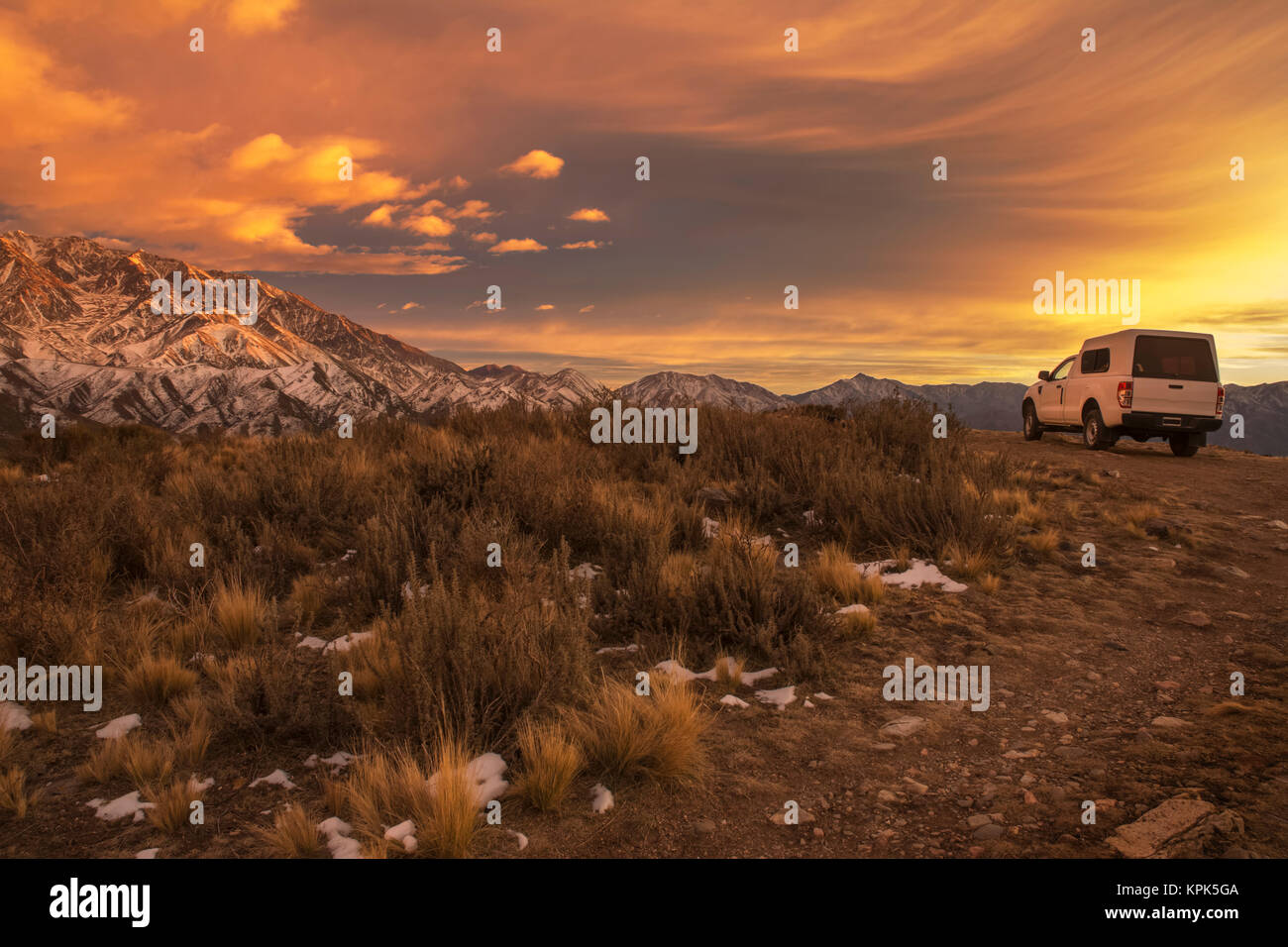 Un petit camion-cabine s'est arrêté dans un paysage désertique avec des montagnes enneigées en arrière-plan.Le lever du soleil illumine les montagnes et les nuages avec bri... Banque D'Images