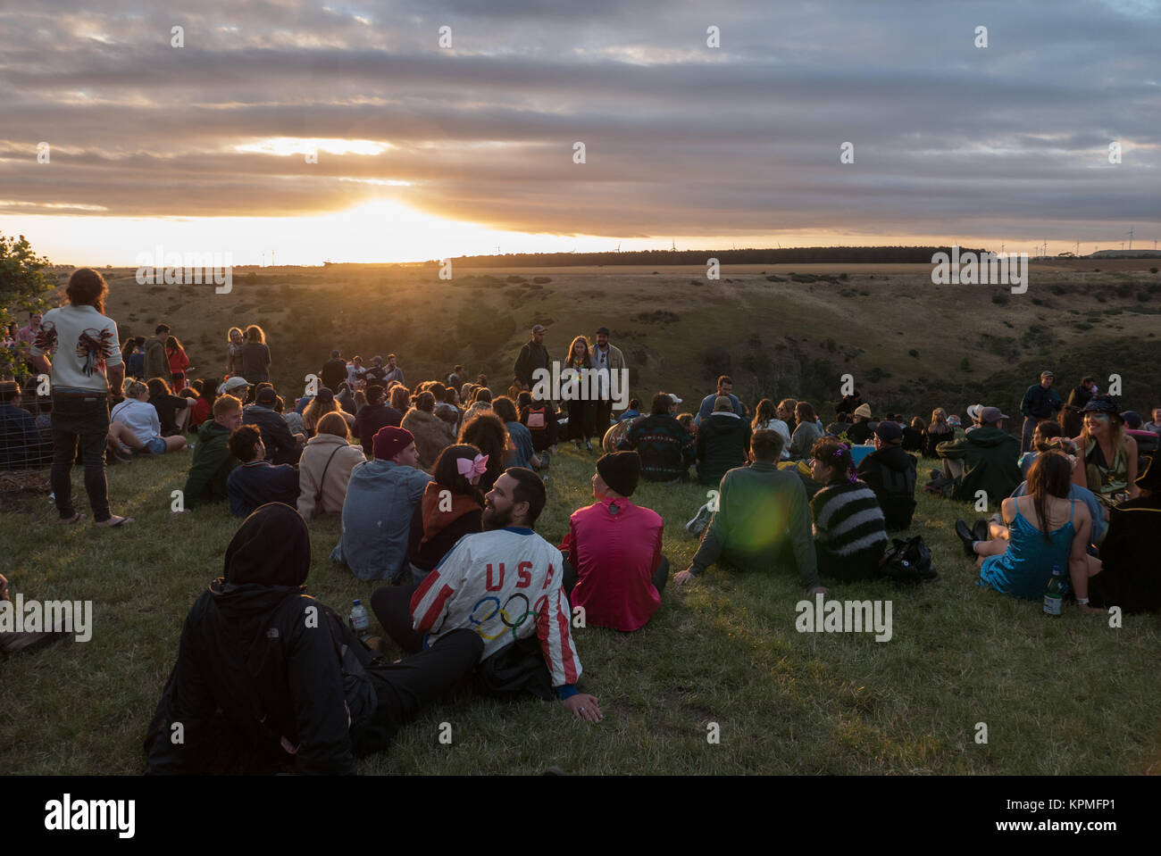 Grand groupe de jeunes, habillés de couleurs vives les festivaliers musique magique de regarder un coucher de soleil d'or à l'horizon avec les éoliennes en arrière-plan. Banque D'Images