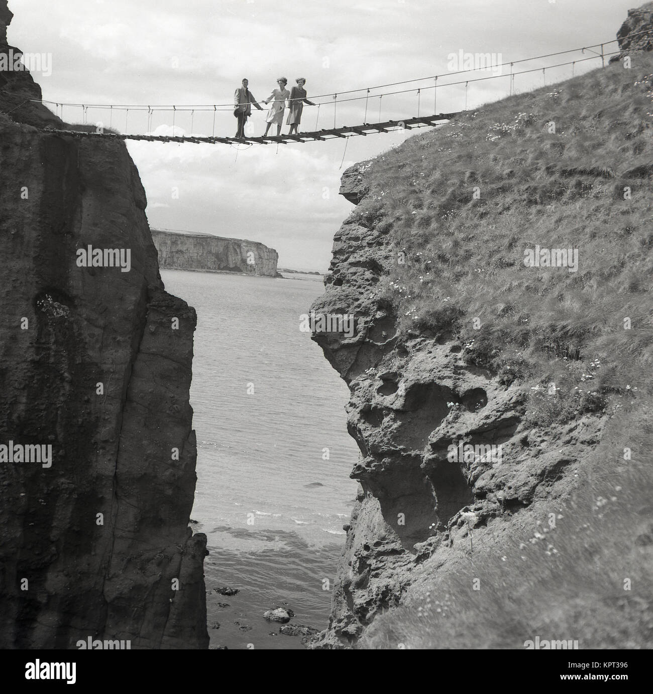 Années 1950, historique, un homme et deux femmes se tenir sur le célèbre pont de corde de Carrick-a-rede, Ballintoy, d'Antrim, en Irlande du Nord. Le pont est à 30 mètres au-dessus les roches ci-dessous. Banque D'Images