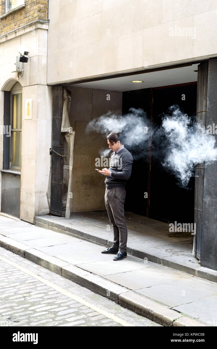 Man in casual vêtements d'affaires se trouve à l'extérieur une entrée du bâtiment à l'aide de son téléphone et le tabagisme avec de grands panaches de fumée Banque D'Images