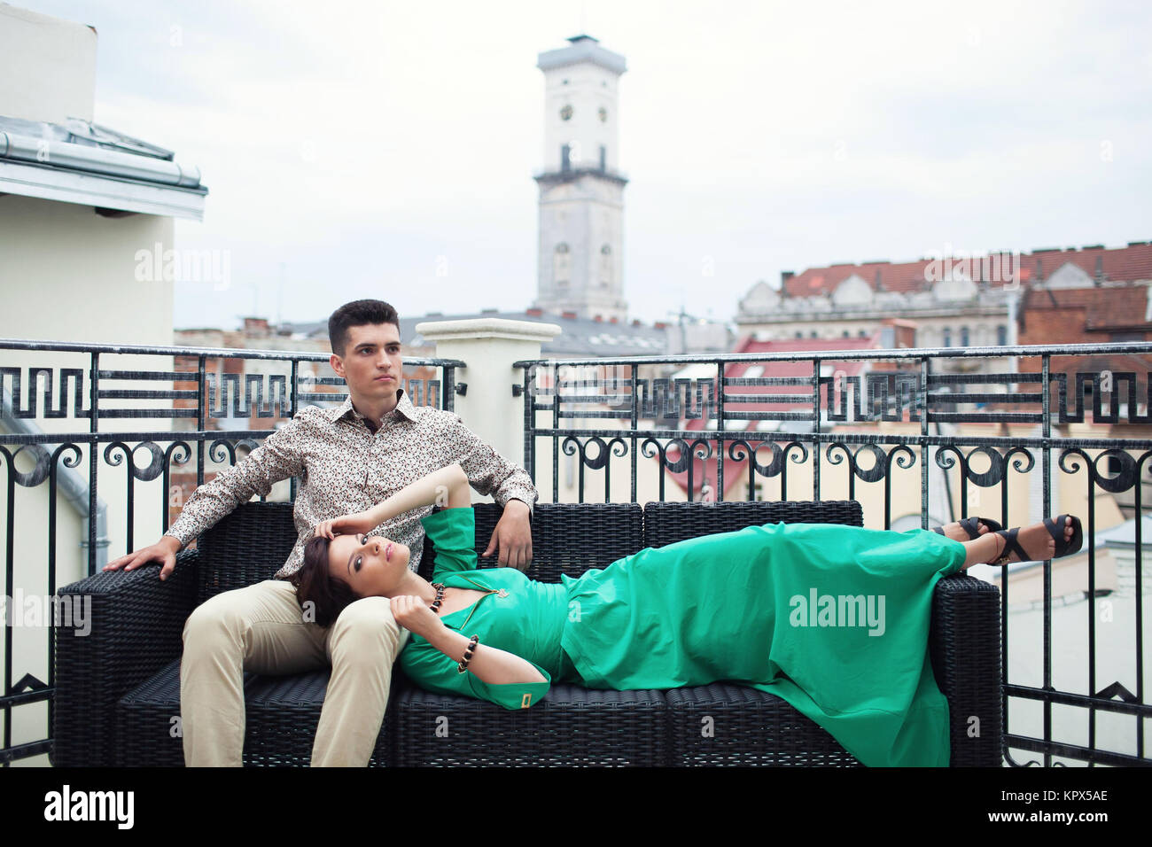 Ð¡ouple détente sur la terrasse balcon avec de beaux paysages sur l'ancienne ville. Brunette guy dans une chemise et le pantalon beige assis sur le canapé avec une fille qui lui mentir à genoux dans une robe verte. Banque D'Images