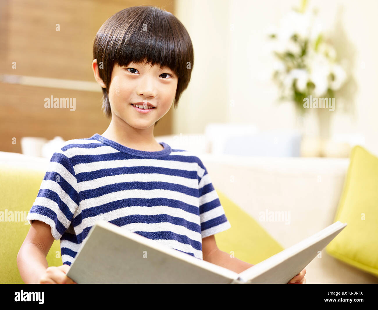 7 ans, petit garçon asiatique assis sur la table tenant un livre à la caméra en souriant. Banque D'Images