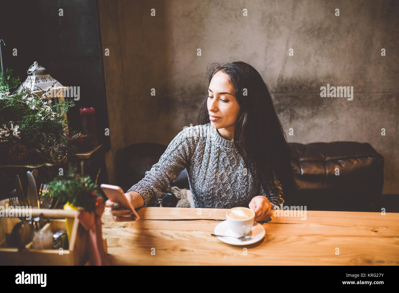 Belle jeune fille utilise, les types de texte sur un téléphone mobile à une table en bois près de la fenêtre et boit du café dans un café décoré avec des décorations de Noël. Vêtu d'un pull en laine tricoté gris Banque D'Images