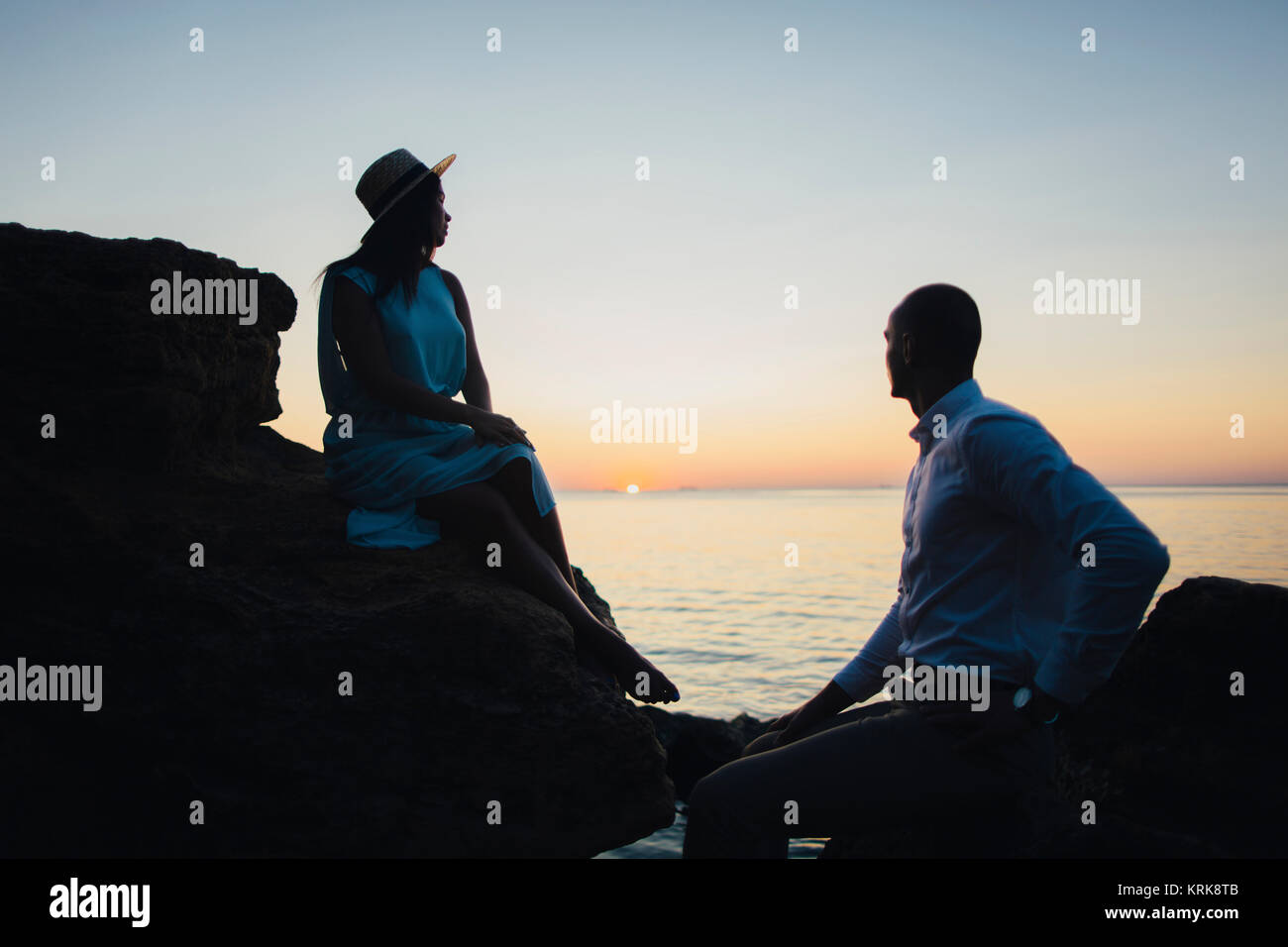 Couple assis sur des rochers près de l'océan au coucher du soleil Banque D'Images