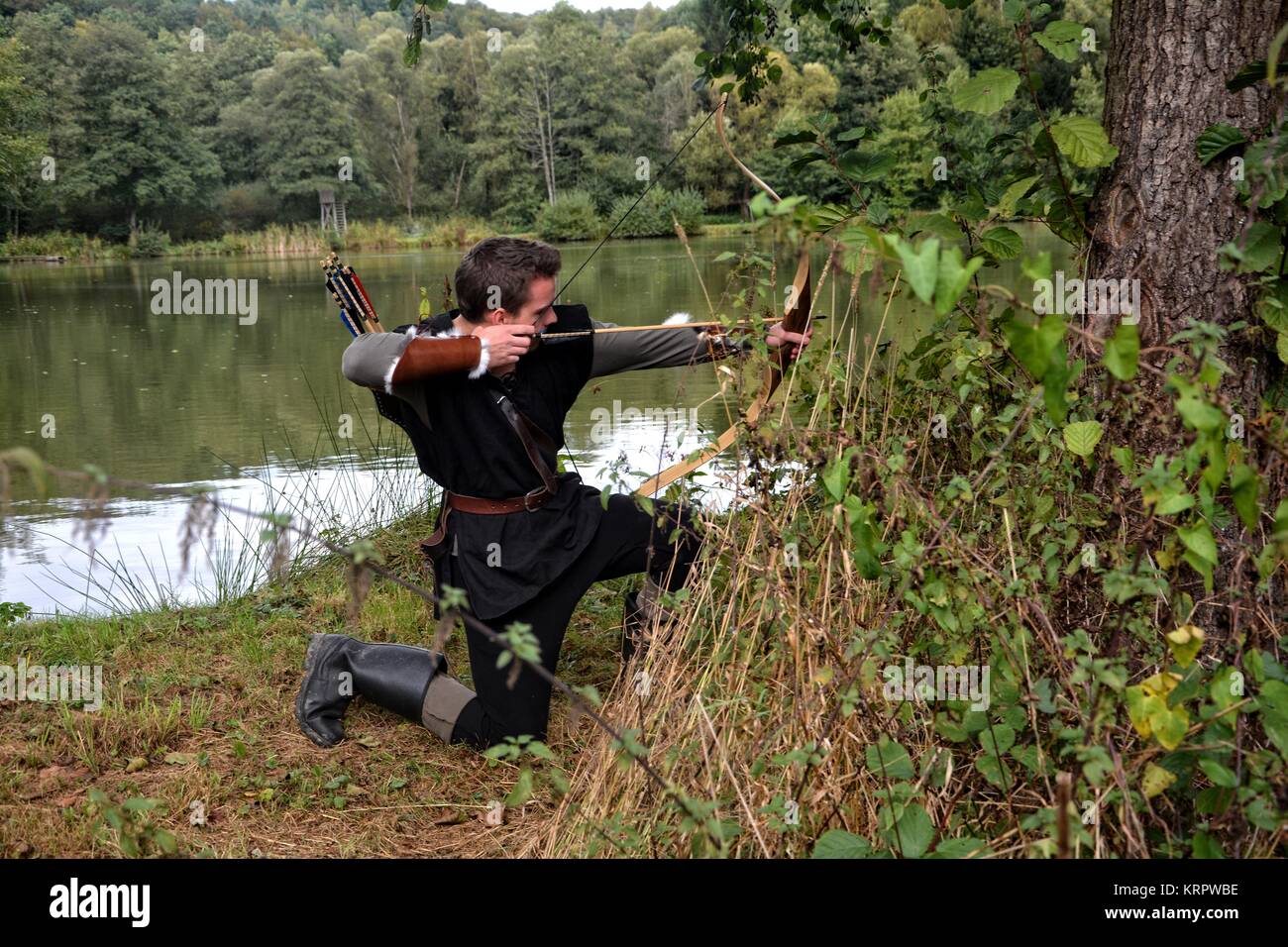 Archer médiéval avec capot noir s'agenouille sur le côté de la terre, visant avec la flèche et l'arc sur le côté Banque D'Images