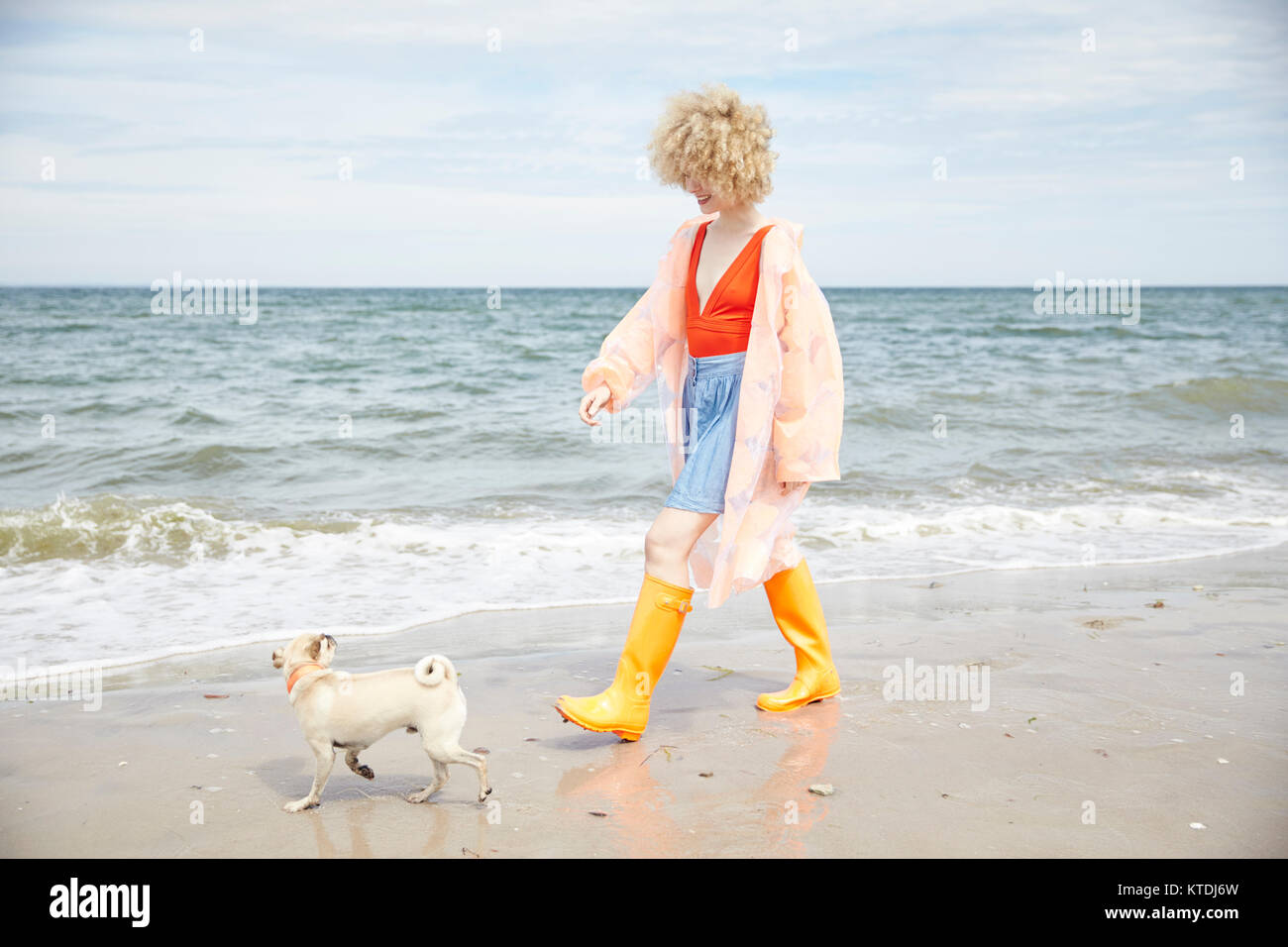 Smiling young woman wearing rain Manteau et bottes Wellington passerelles va avec son chien au bord de mer Banque D'Images