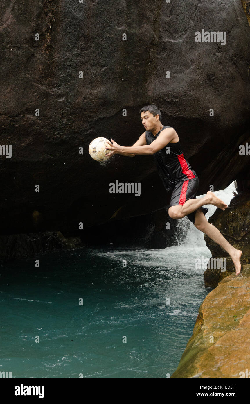 Jeune adulte Jeune Indien sauter d'une petite falaise en frais de bleu piscine de l'eau, tenant une balle sur un Nagarmadi Chendia, chutes d'eau, Karnataka, Inde. Banque D'Images