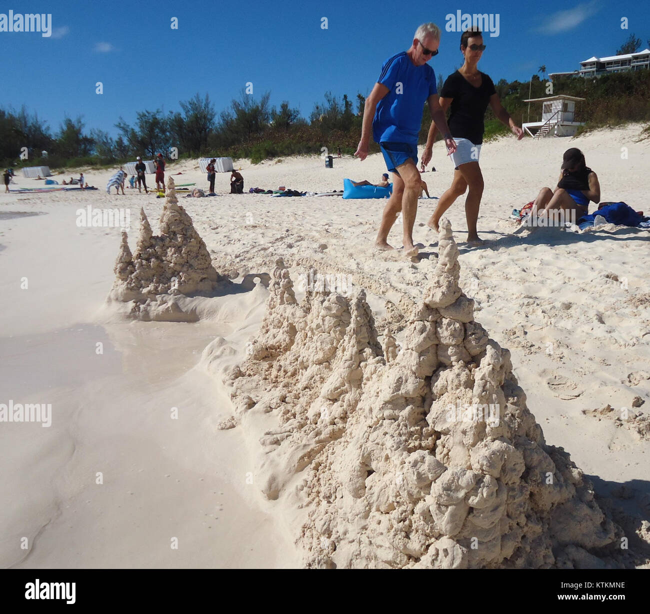 Cette image des Bermudes (Royaume-Uni) montre un château de sable sur la plage, avec un couple marchant le long du rivage. La scène balnéaire met en valeur la beauté du littoral des Bermudes, avec des eaux claires et des plages de sable blanc. Banque D'Images