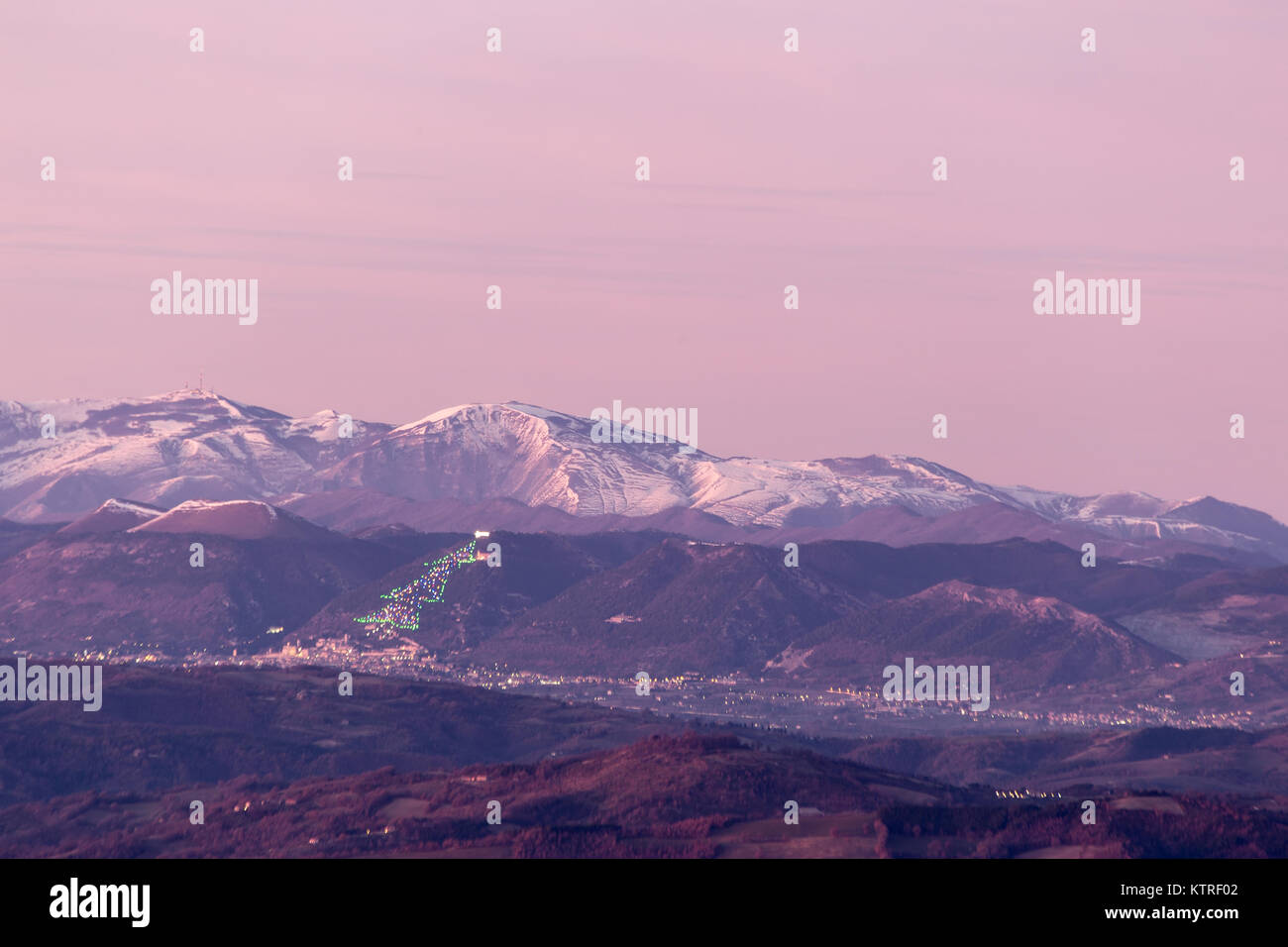 La vallée de l'Ombrie en hiver, avec une vue de la ville de Gubbio, avec de grands arbres de Noël éclairé sur le flanc de la montagne et Monte Cucco recouvert de neige Banque D'Images