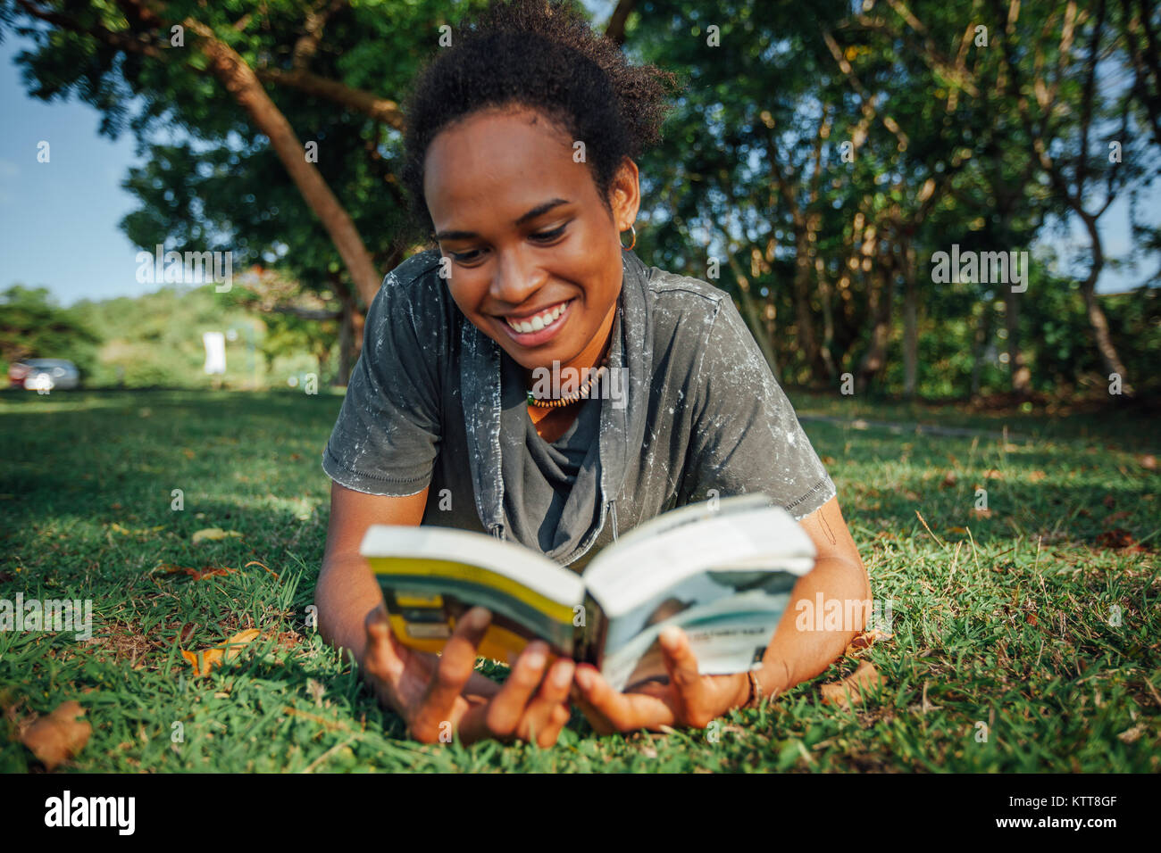 Belle jeune femme avec coiffure afro et livre dans ses mains, allongé sur l'herbe dans le parc aux beaux jours d'été Banque D'Images