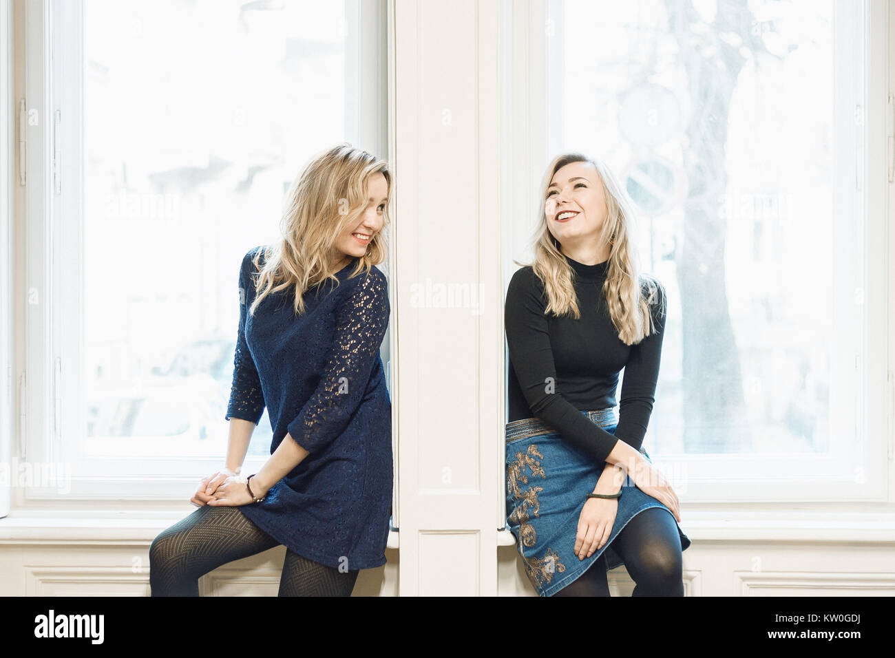 Deux blonde female friends chatting et rire tout en restant assis par le mur entre les deux fenêtres. Briser la barrière entre les personnes concept Banque D'Images