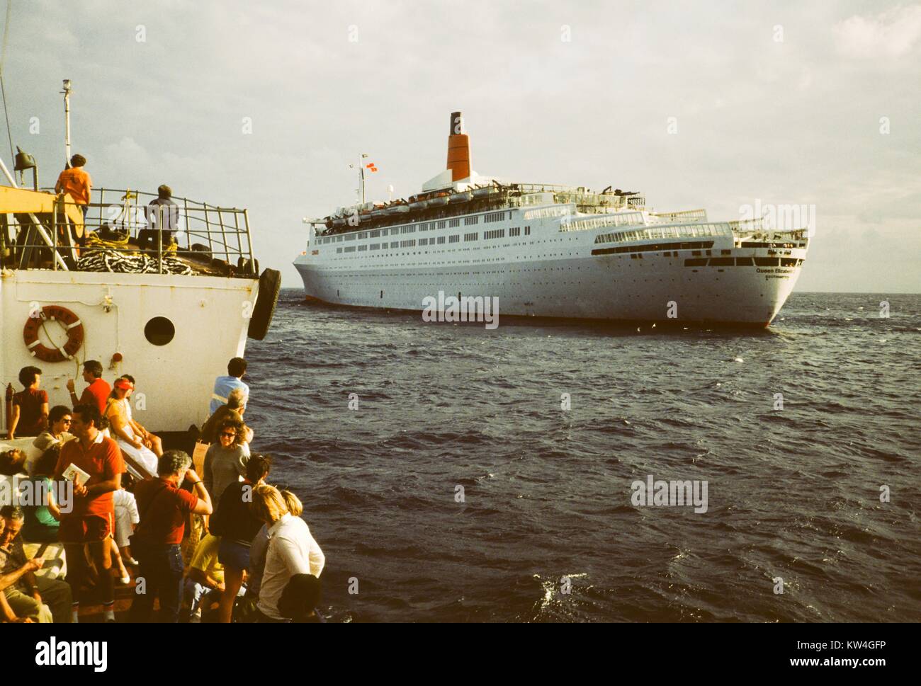 Les touristes se tiennent sur un petit bateau à l'approche de la ligne Cunard Queen Elizabeth 2 bateau de croisière en mer, 1975. Banque D'Images