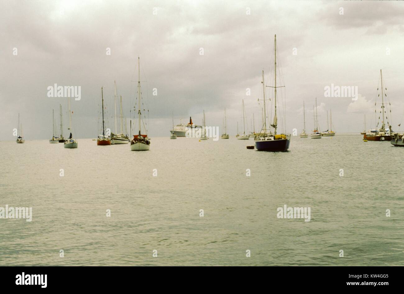 La ligne Cunard Queen Elizabeth 2 bateau de croisière est visible dans la distance à travers un plan d'eau, avec de petits voiliers dans le premier plan, sur un jour nuageux, 1975. Banque D'Images