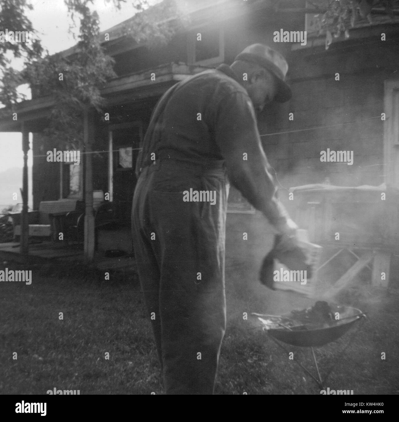 Homme mûr se pencha plus près d'un petit barbecue à l'extérieur de sa maison de banlieue, le coulage des briquettes de charbon sur le grill comme fumée entoure, 1950. Banque D'Images