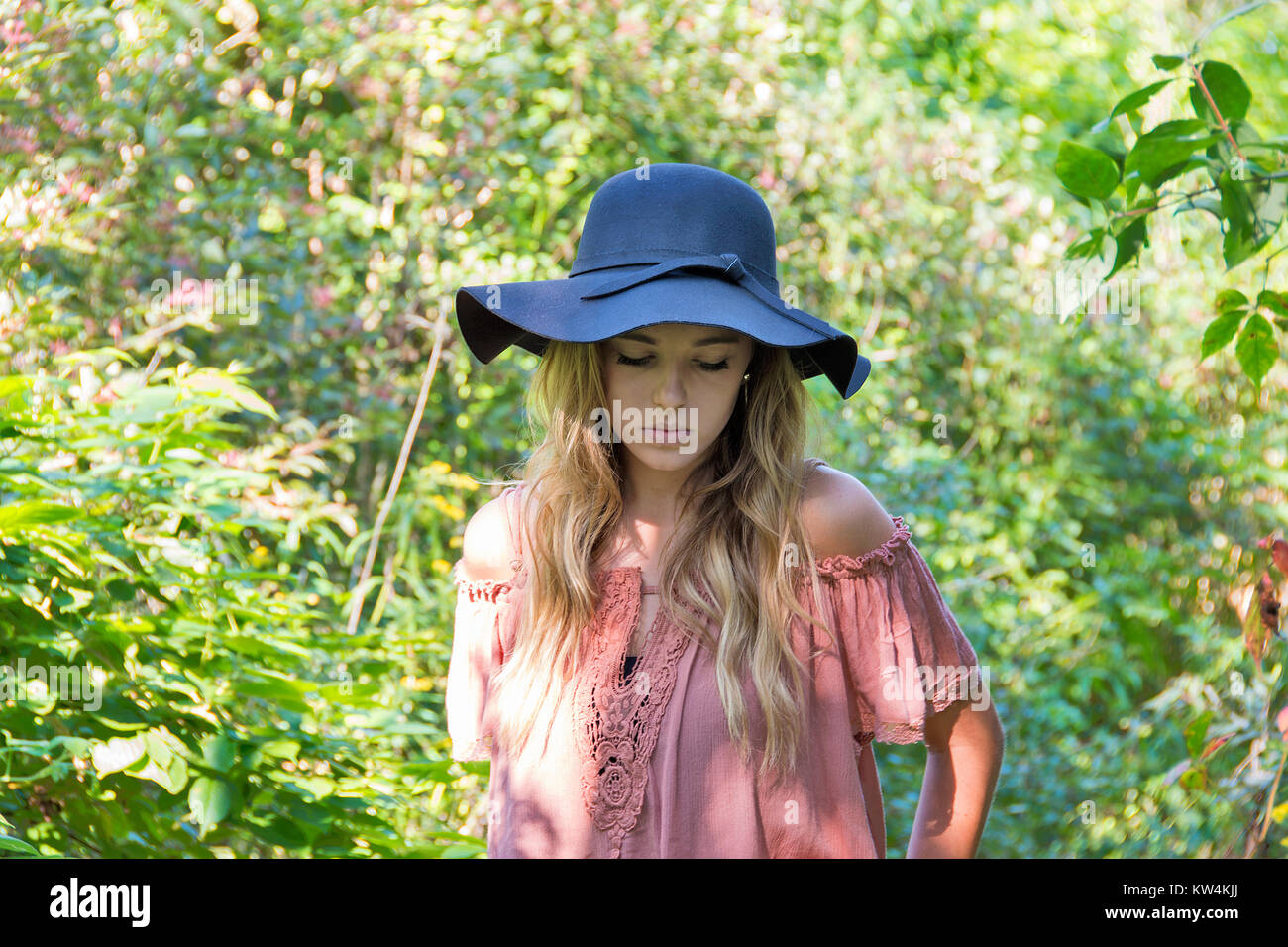 Caucasian teenage girl in sunlit summer woods avec expression grave portant chapeau noir et de l'épaule chemisier. Banque D'Images