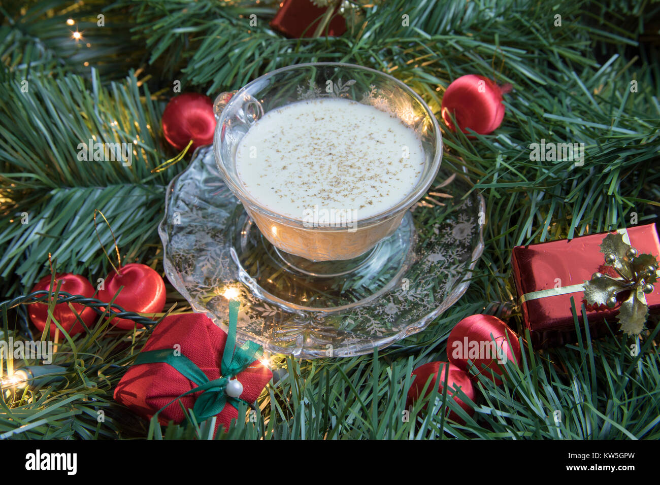 Close up of crystal Cup and Saucer with egg nog saupoudrée de muscade dans Garland avec les lumières et les red ball des ornements et des paquets. Banque D'Images