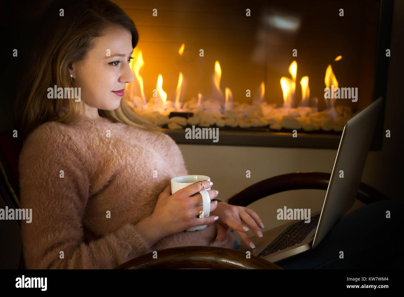 Jeune femme, 20 ans, avoir une tasse de café à l'aide d'un ordinateur portable assis sur une chaise à bascule près d'une cheminée, de détente à la maison. Banque D'Images