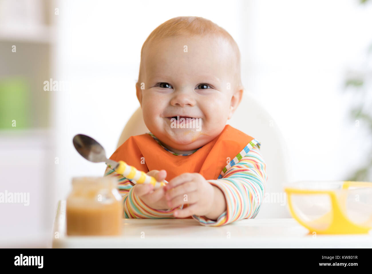 Cheerful baby enfant mange la nourriture elle-même avec une cuillère. Portrait of happy kid garçon en chaise haute. Banque D'Images