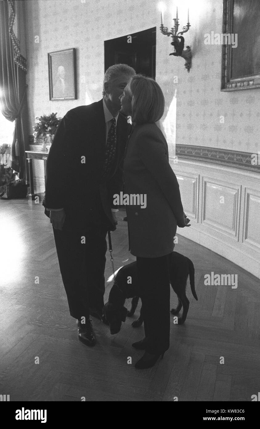 À l'intérieur d'une salle à manger de la Maison Blanche, le président Clinton se trouve à côté de la première dame Hillary Rodham Clinton, tous deux portant des costumes de pantalon, smiling while chuchotant à son oreille, tenant une laisse pour premier animal, Buddy le chien, un labrador brun moka, qui est à leurs pieds de la capture d'un parfum sur le plancher, Washington, District de Columbia, le 18 décembre 1997. Banque D'Images