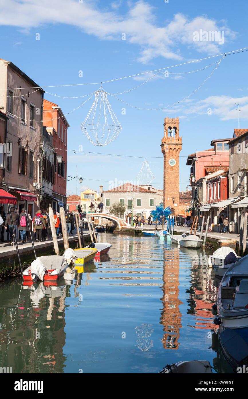 L'île de Murano, Venise, Italie, la Tour de l'horloge dans le Campo San Stefano reflétée dans Canal Vetrai avec bateaux colorés et touristes en hiver Banque D'Images
