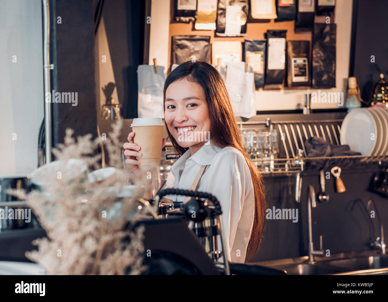 Asian female barista de porter un tablier jean holding cup à emporter bar comptoir machine avec smile face,cafe service concept.faire le café Banque D'Images