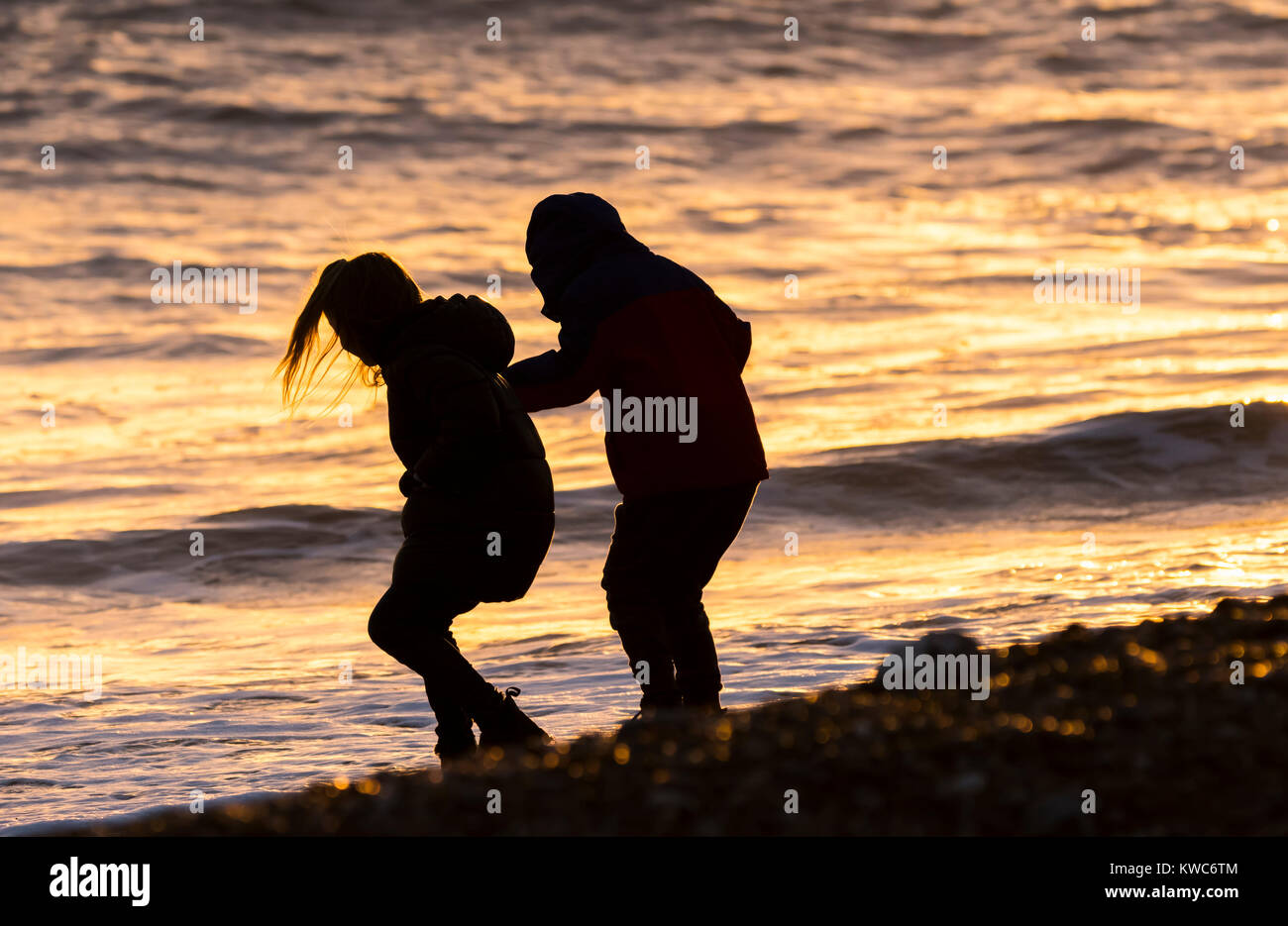 Silhouette de jeune mâle et femelle childten jouant sur la plage au coucher du soleil et se reflète dans la mer en hiver. Banque D'Images
