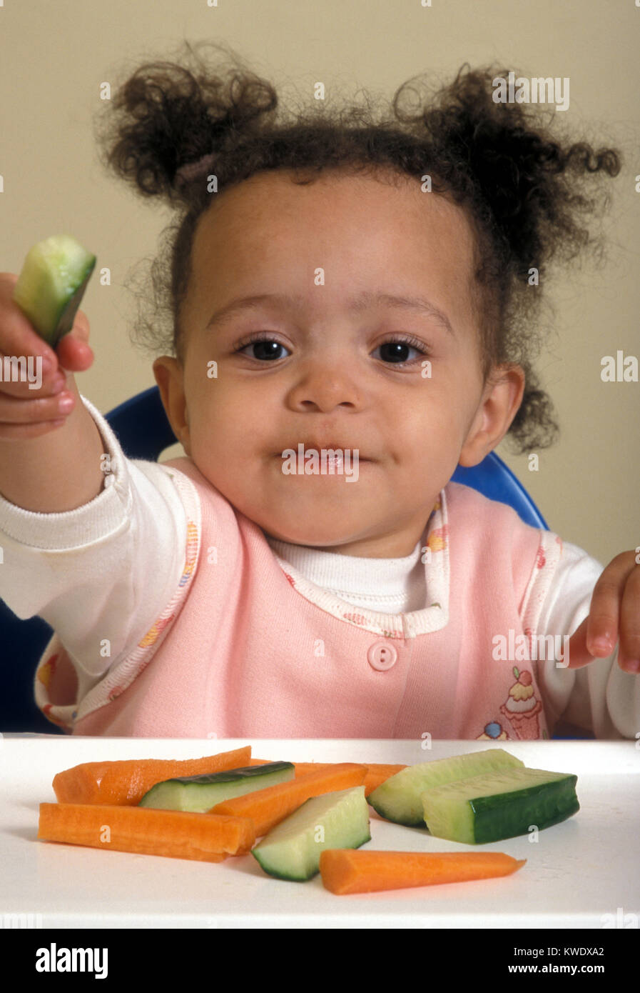 Mixed Race bébé dans une chaise haute de manger des bouchées de carotte et concombre Banque D'Images