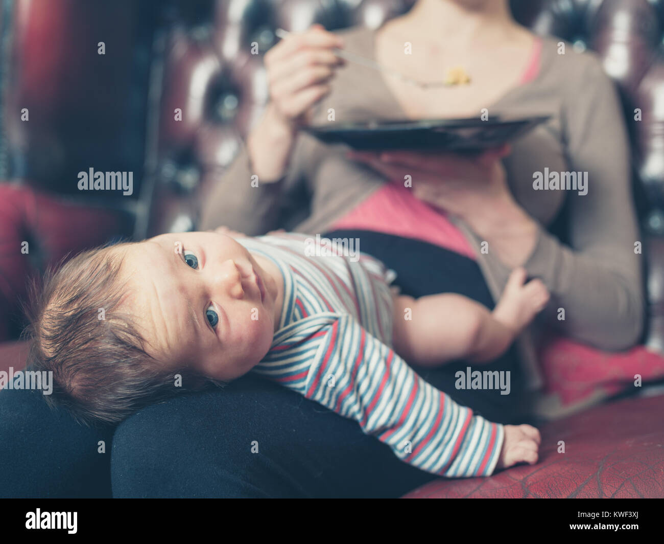 Une jeune mère est assise sur un canapé et essaie de manger son dîner avec son bébé sur ses genoux Banque D'Images