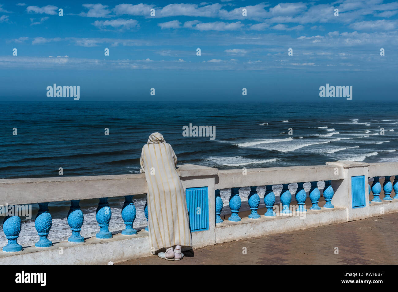 En Djellaba marocaine solitaire regarde vers la mer, appuyé contre blue balastrade. Banque D'Images
