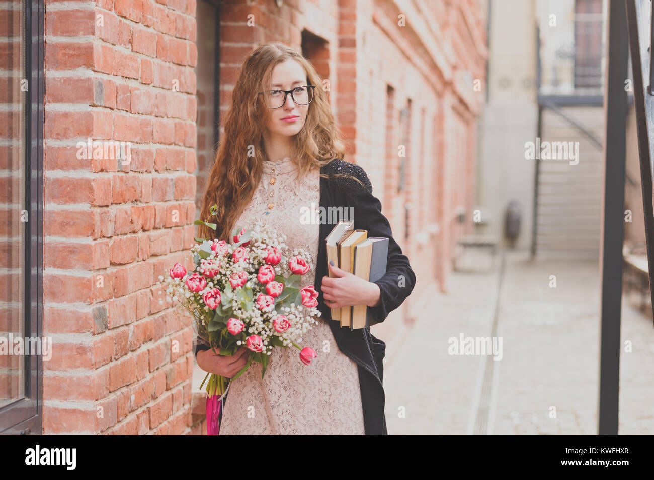 Une femme aux cheveux bouclés portant des lunettes est maintenant un bouquet de tulipes et de colis de livres contre le bâtiment en briques rouges Banque D'Images