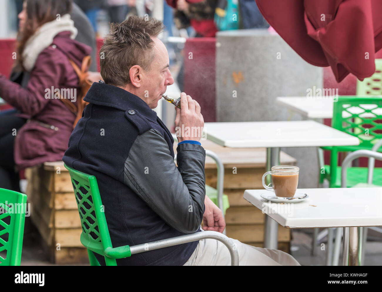 Homme caucasien assis dans un café à l'extérieur en hiver sur une cigarette électronique en Angleterre, au Royaume-Uni. Banque D'Images