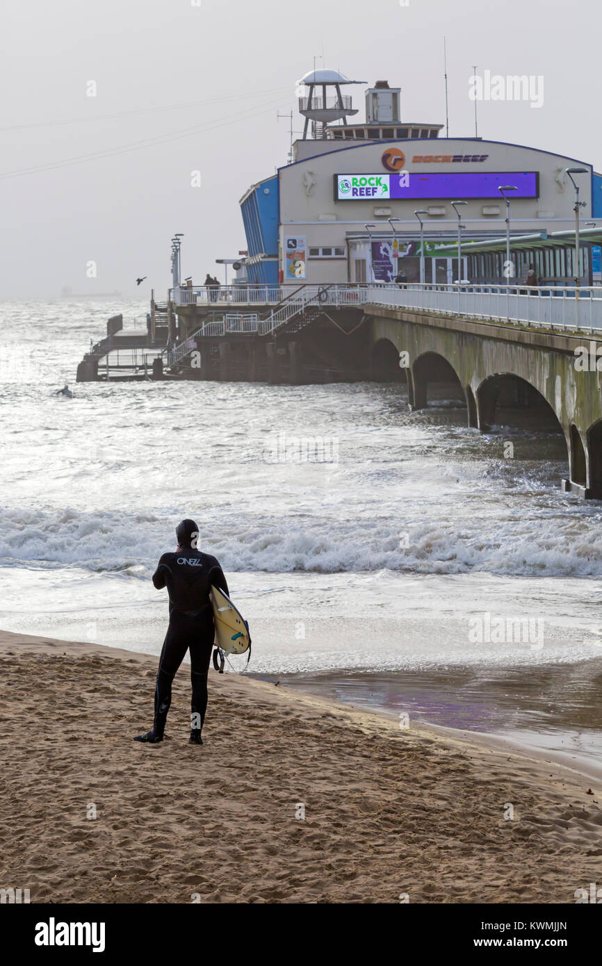 Bournemouth, Dorset, England UK. 4 janvier, 2018. Météo France : surfer debout sur la plage près de la jetée à la mer comme les surfeurs profiter du surf sur un jour de vent à la plage de Bournemouth, que les surfeurs profiter de la vent et de grosses vagues. Banque D'Images