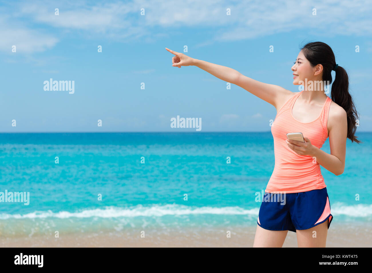 Cheerful attractive female traveler wearing sportswear standing on beach en utilisant smartphone et le partage à distance de pointage célèbre paysage. Banque D'Images