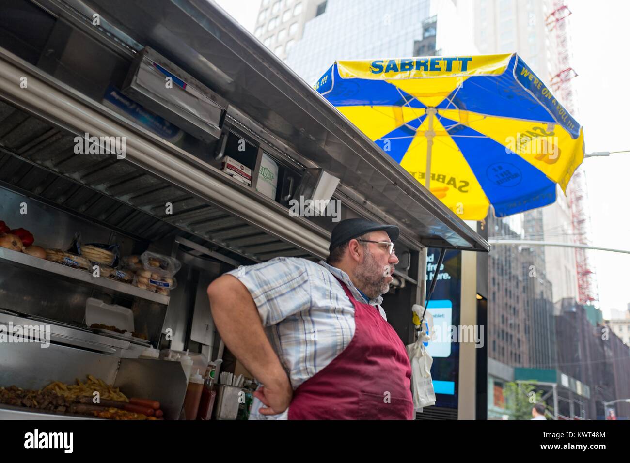 Un fournisseur d'aliments de rue portant un tablier, se distingue avec son panier sur 60th Street à Manhattan, New York City, New York, avec des hot-dogs et autres aliments visible dans le panier et un parapluie bleu et jaune Sabrett visibles au-dessus, le 14 septembre 2017. Banque D'Images