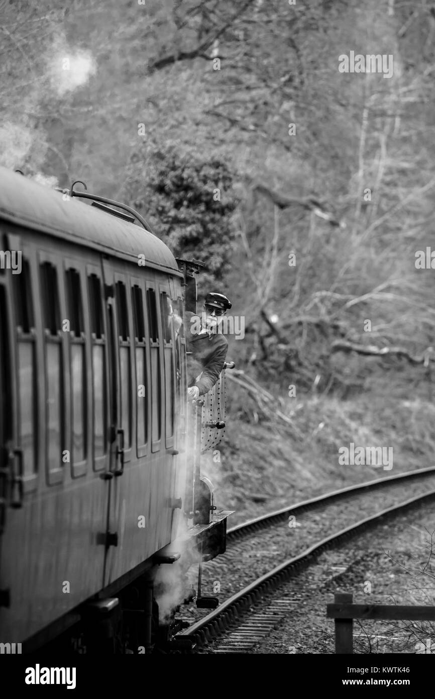 Portrait, tourné en noir et blanc du moteur incliné vers l'extérieur de la cabine du conducteur de train à vapeur britannique regarde ses passagers à bord de l'vintage railway carriage derrière. Banque D'Images