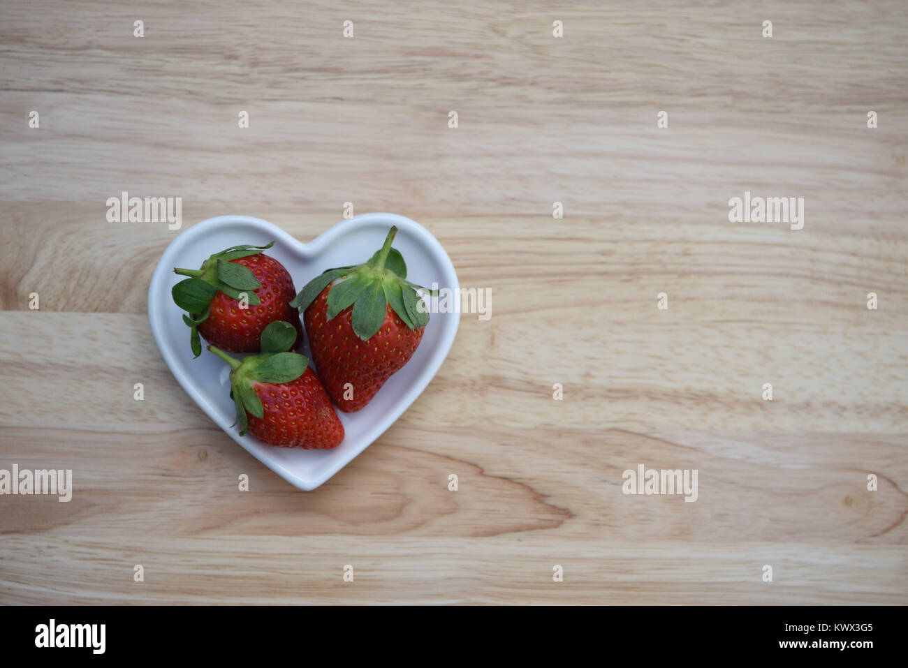 Photographie image alimentaire des fruits frais avec trois rouges fraises avec feuille verte haut placé dans un plat en forme de coeur amour blanc sur fond de bois Banque D'Images