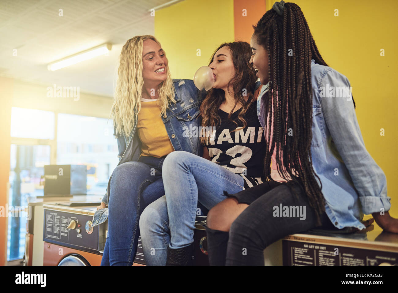 Laughing young female friends sitting on lave-linge dans une laverie automatique et faire des bulles avec un chewing-gum Banque D'Images