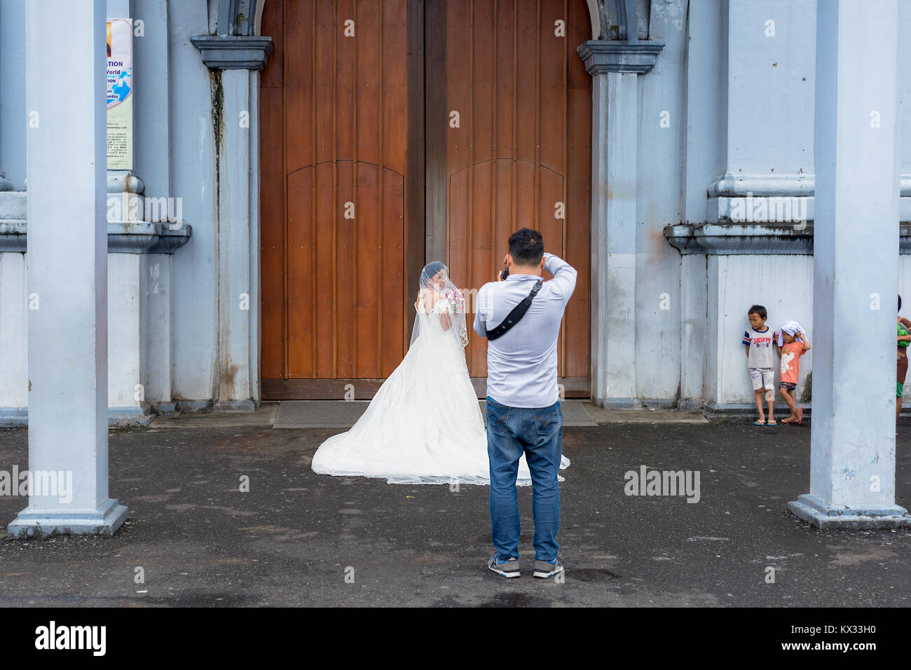 Un photographe de mariage en prenant la dernière photo avant la mariée dans sa robe de mariée blanche entre dans l'église tandis que les enfants regardent locales appauvries Banque D'Images