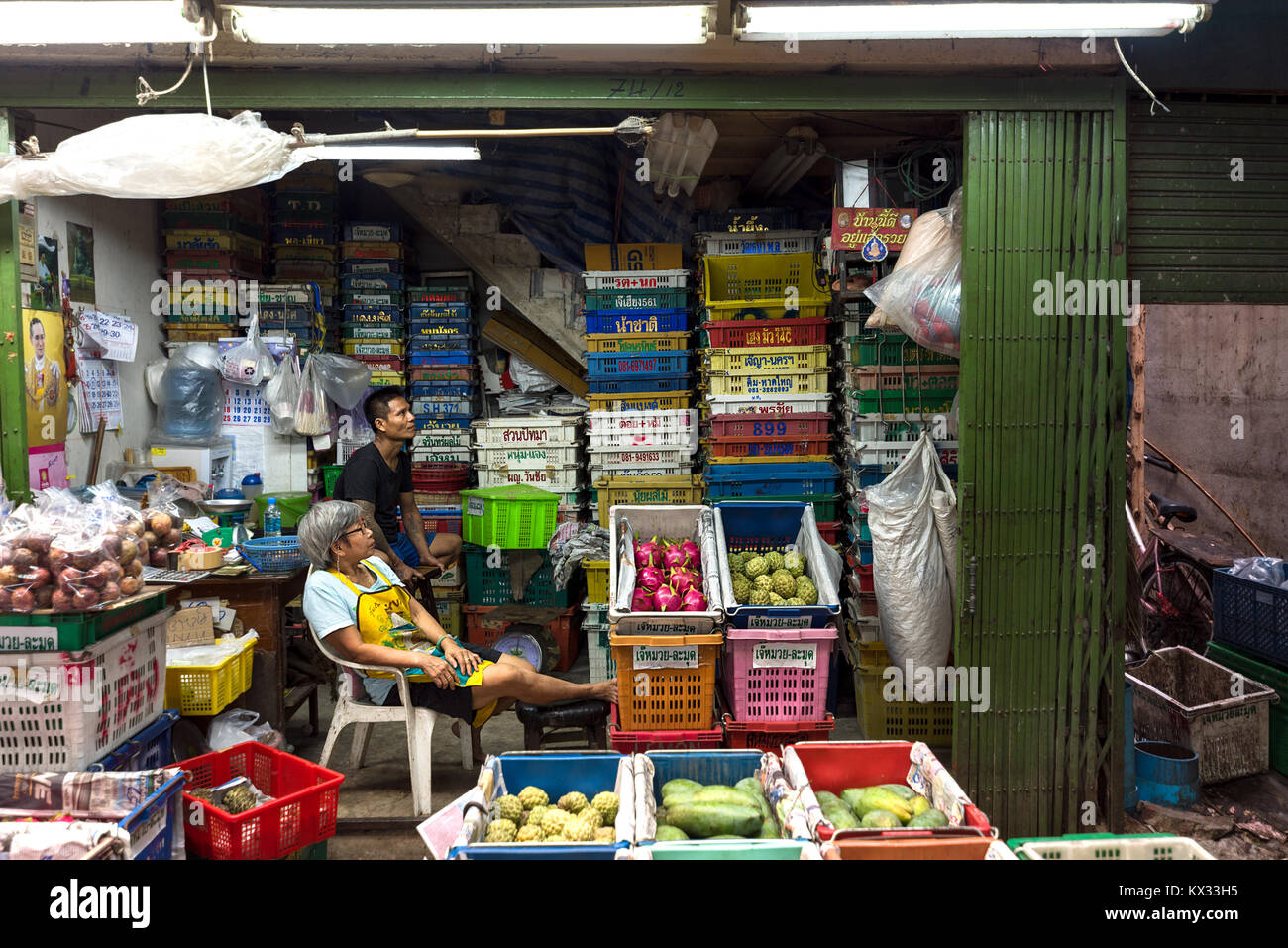 Marché de gros de fruits asiatiques deux détenteurs de décrochage assister à leur stand à regarder la télévision entourée de fruits exotiques. Banque D'Images