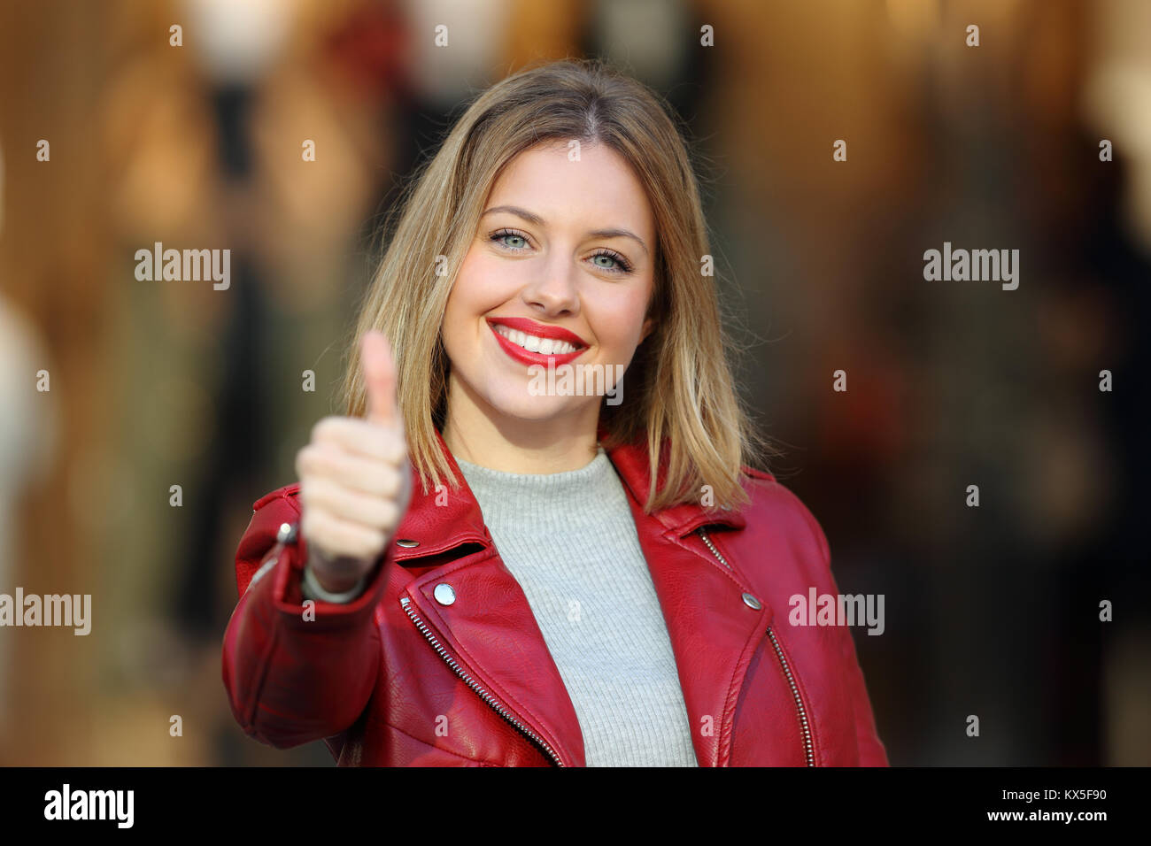 Vue avant portrait of a fashion girl with thumb up portant une veste rouge vous regarde dans la rue avec une devanture en arrière-plan Banque D'Images