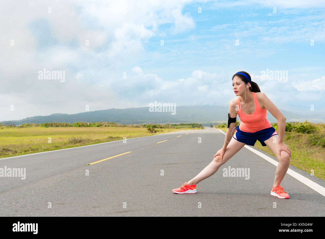 Young pretty female jogger portant des vêtements de sport avec appareil standing on country road stretching jambes réchauffer corps prêt à former d'entraînement. Banque D'Images