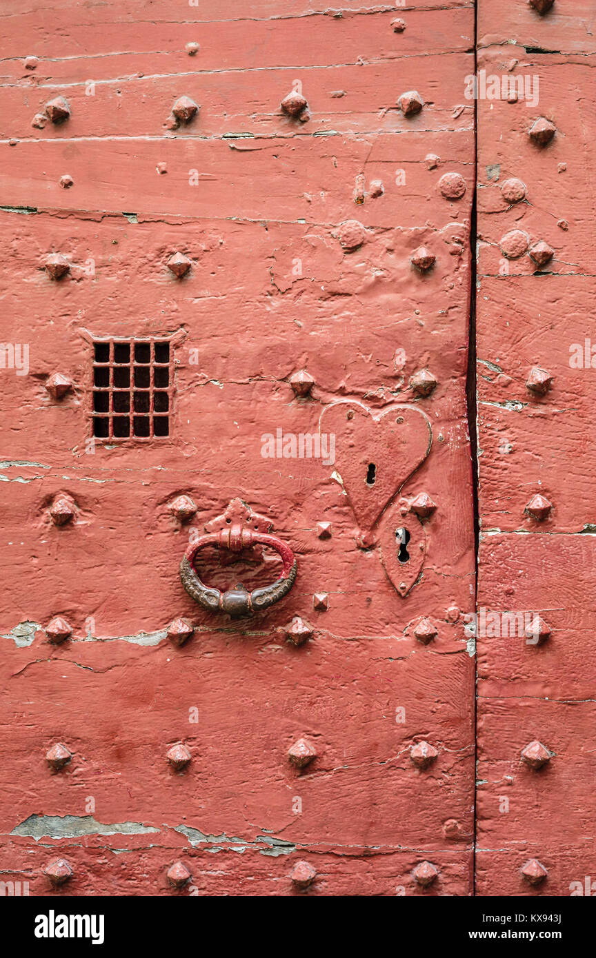 Vue rapprochée d'une ancienne porte rouge de style médiéval avec rivets métalliques, plaque de serrure en forme de coeur, fonte knocker et treillis métallique spyhole. Banque D'Images