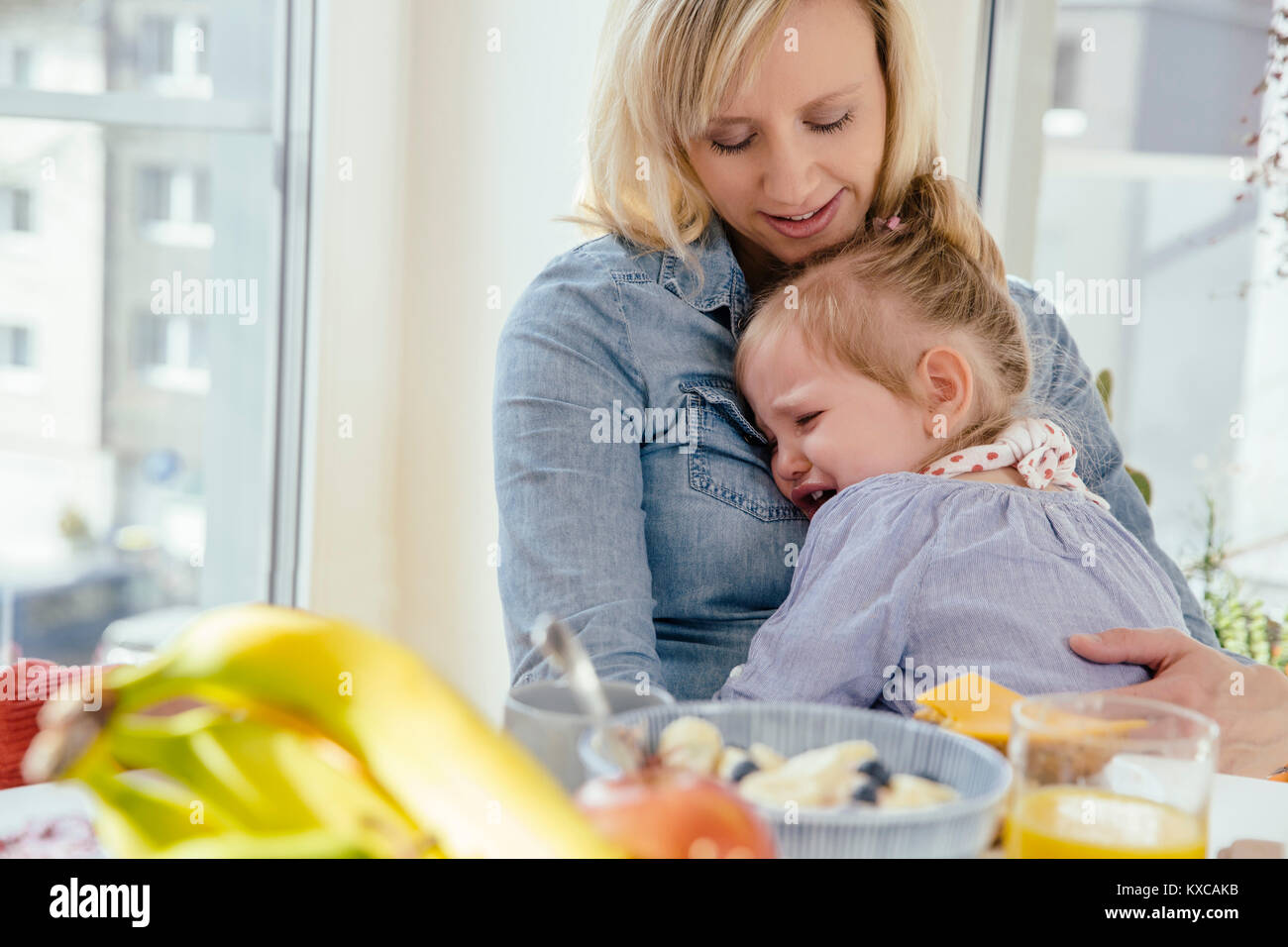 Petite fille pleurer dans les bras de mère à table de petit déjeuner Banque D'Images