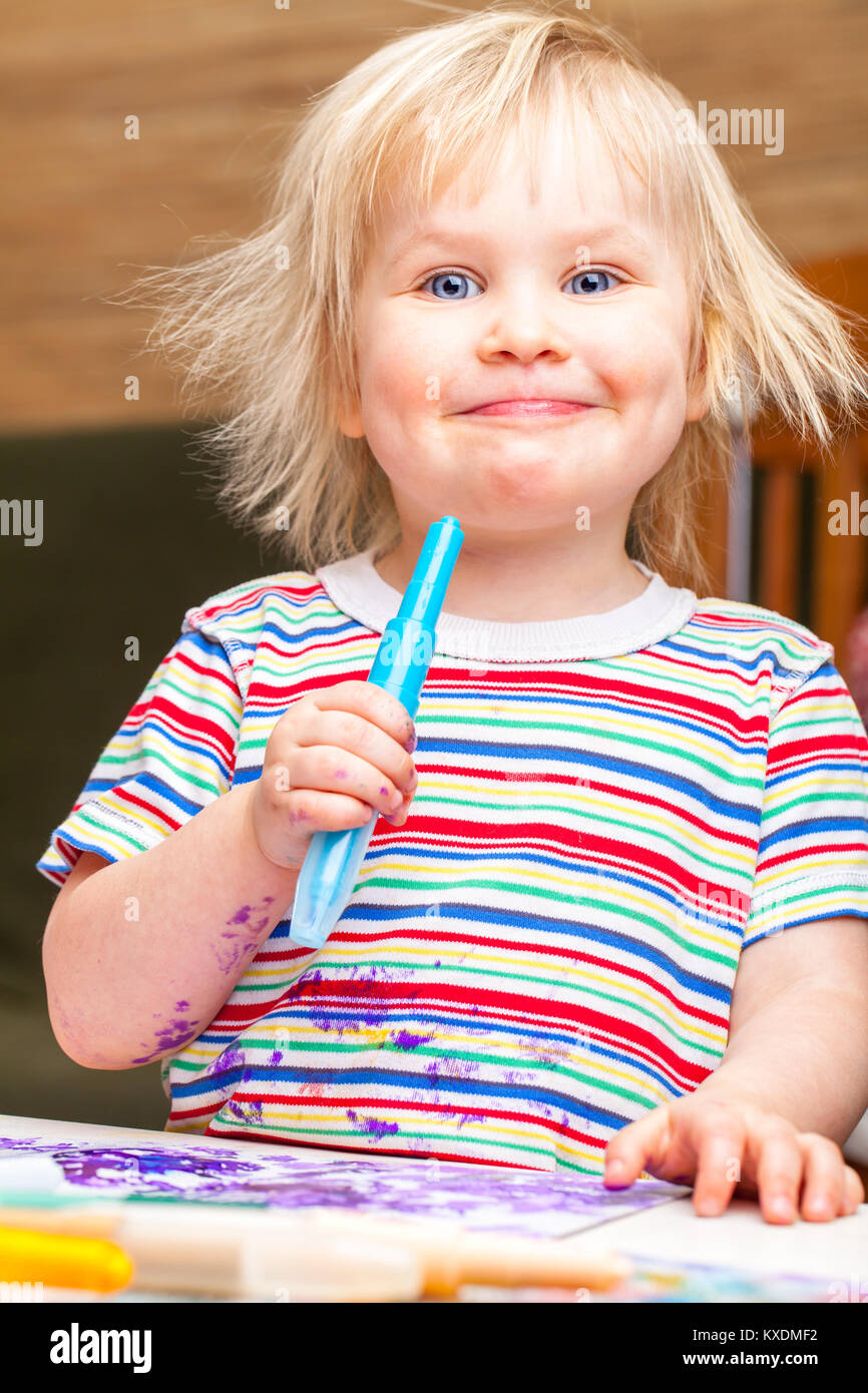 Portrait of happy little Girl with messy cheveux blonds portant des vitraux de couleur tshirt assis à une table à dessin avec des marqueurs de couleur Banque D'Images