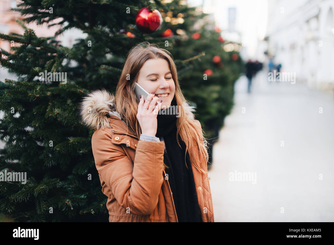 Happy girl talk sur son téléphone et regardez autour de vous avec impression sourire sur son visage. L'heure d'hiver avec arbre sur la rue. Banque D'Images