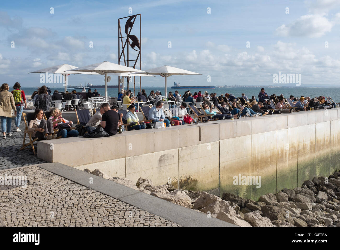 Les gens boire et manger à l'extérieur d'un café sur la promenade au bord de l'eau sur Ribeira das Naus, Lisbonne, Portugal à la fin de l'automne Sunshine Banque D'Images
