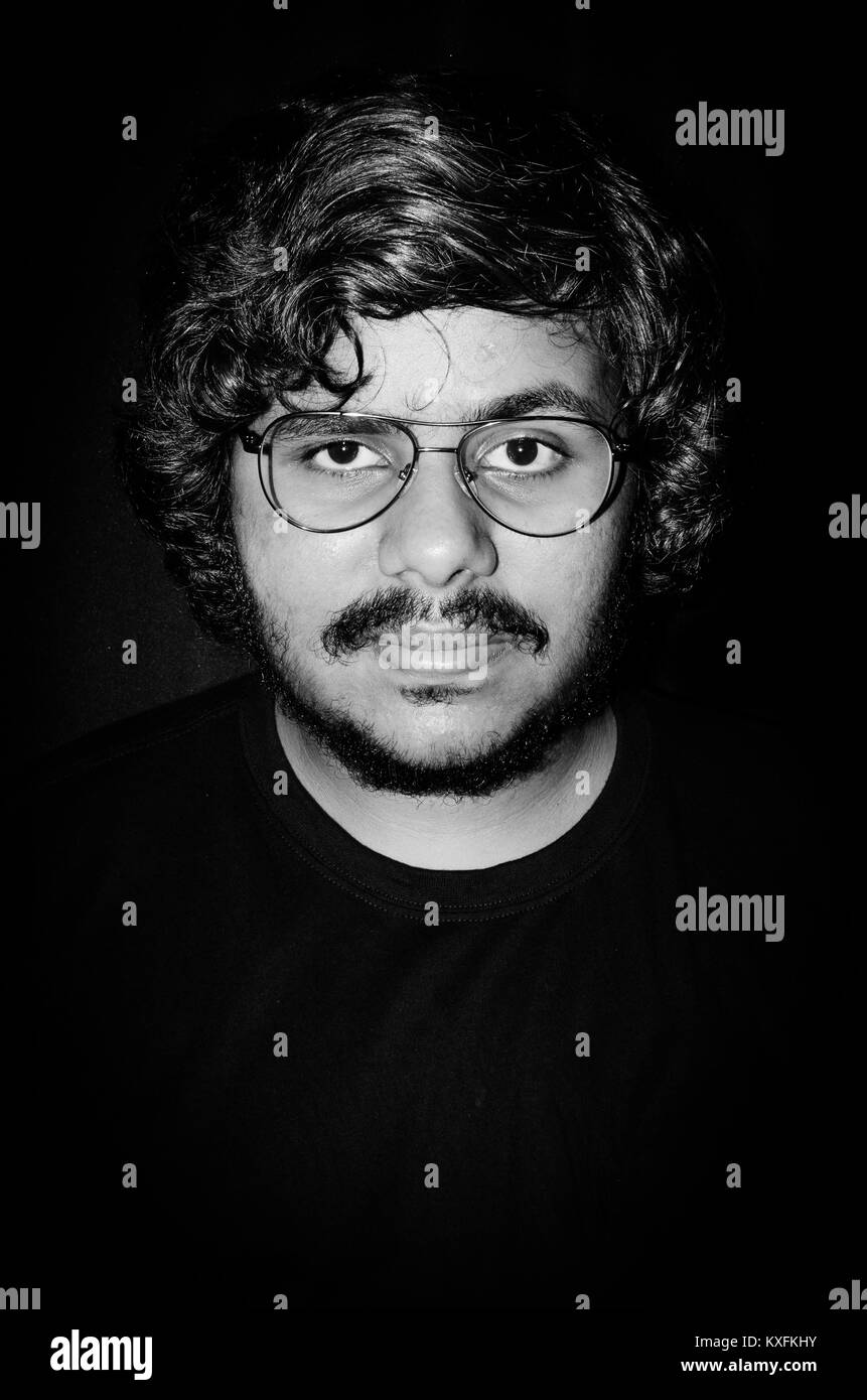 Moody Dark Portrait de jeune homme à lunettes Escobar regardant droit dans la caméra sur fond noir. Banque D'Images